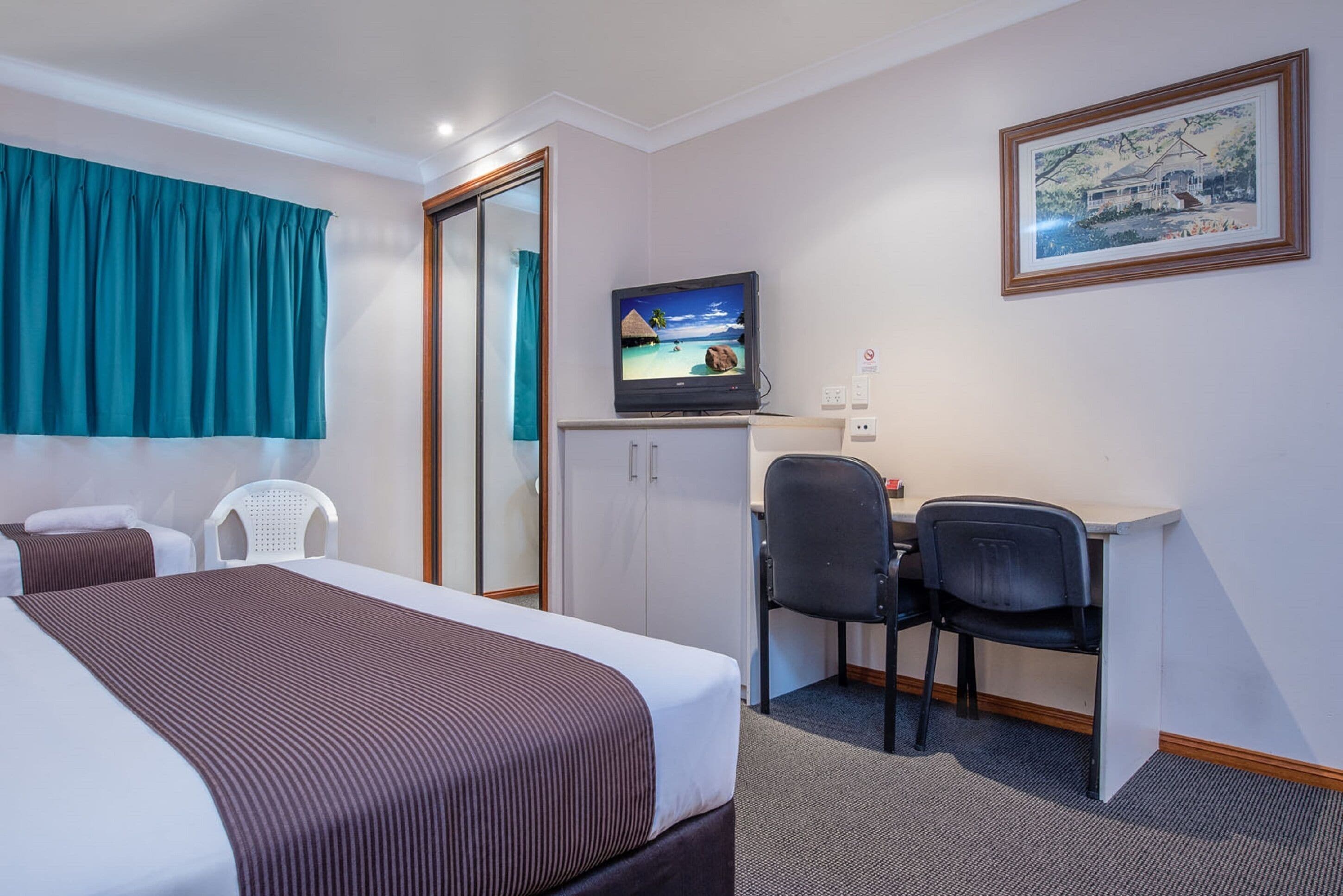Habitación de hotel con cama, escritorio con dos sillas, televisor, armario y un cuadro en la pared.