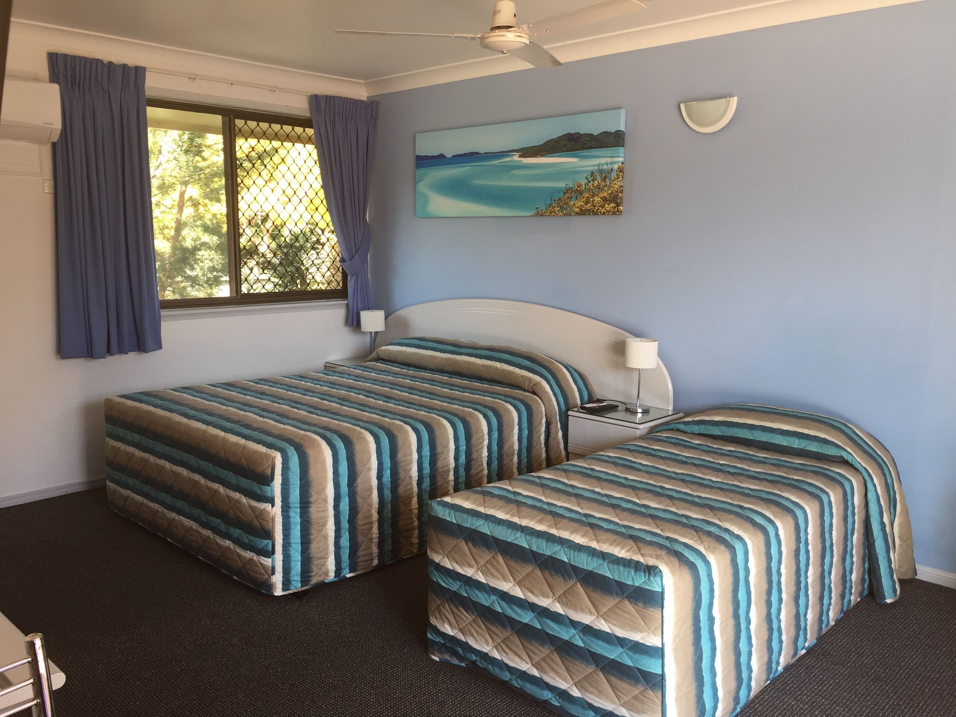A room with two beds, a ceiling fan, a painting of a coastal scene, and a window with curtains.