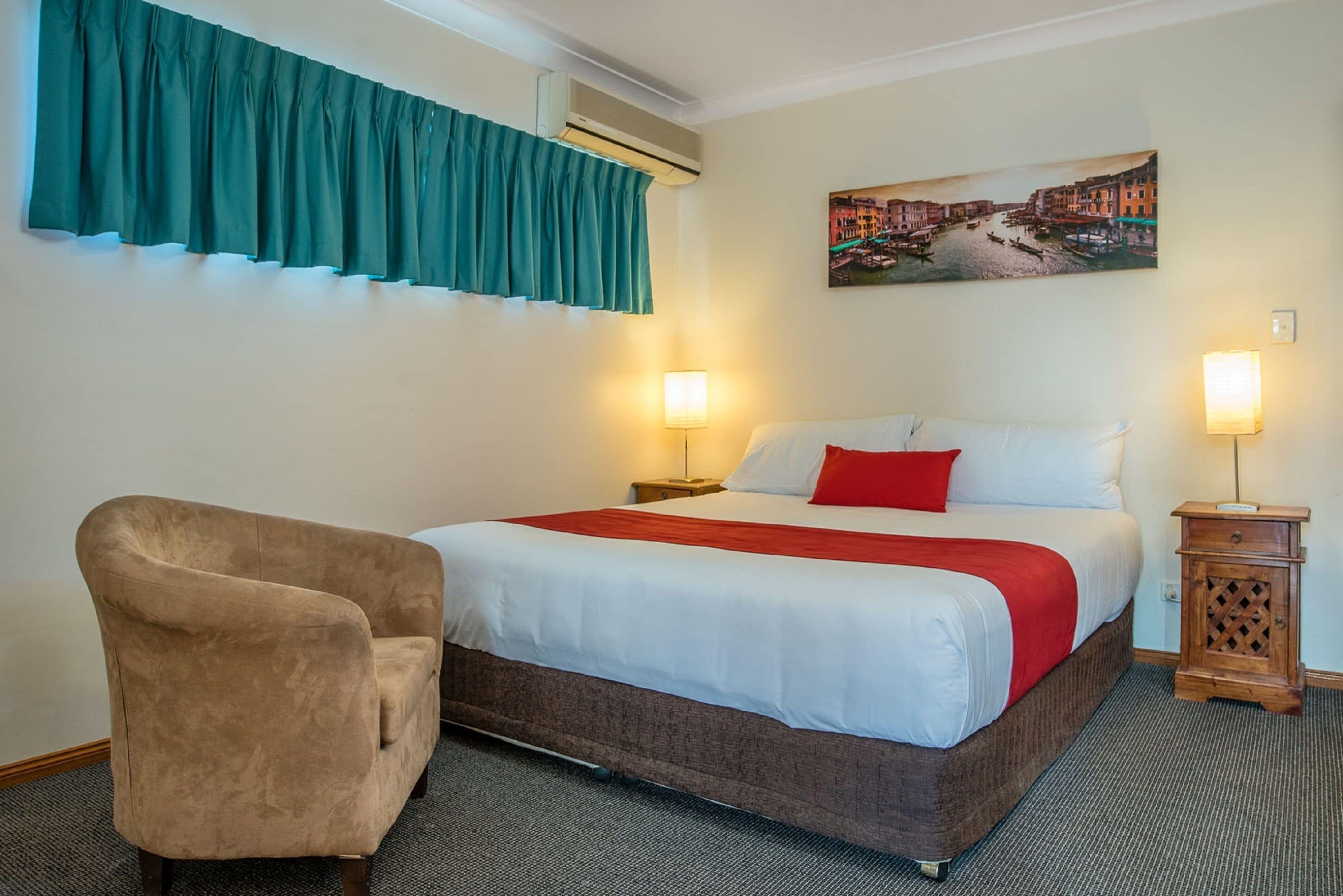 A hotel room with a bed, a chair, a nightstand, and a wall-mounted air conditioner.