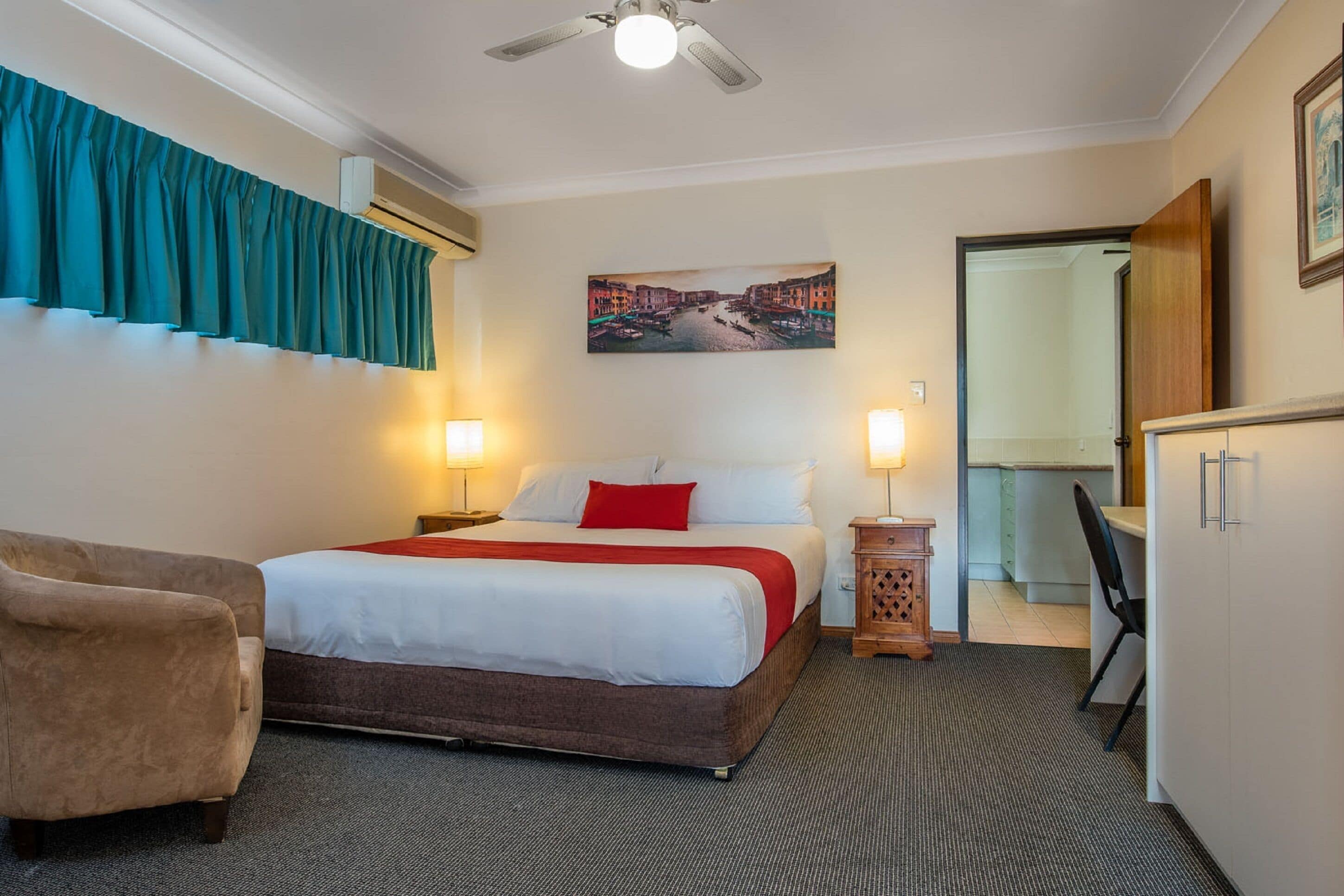 Une chambre d’hôtel avec un lit, un bureau, une chaise, un ventilateur de plafond et un tableau au mur.