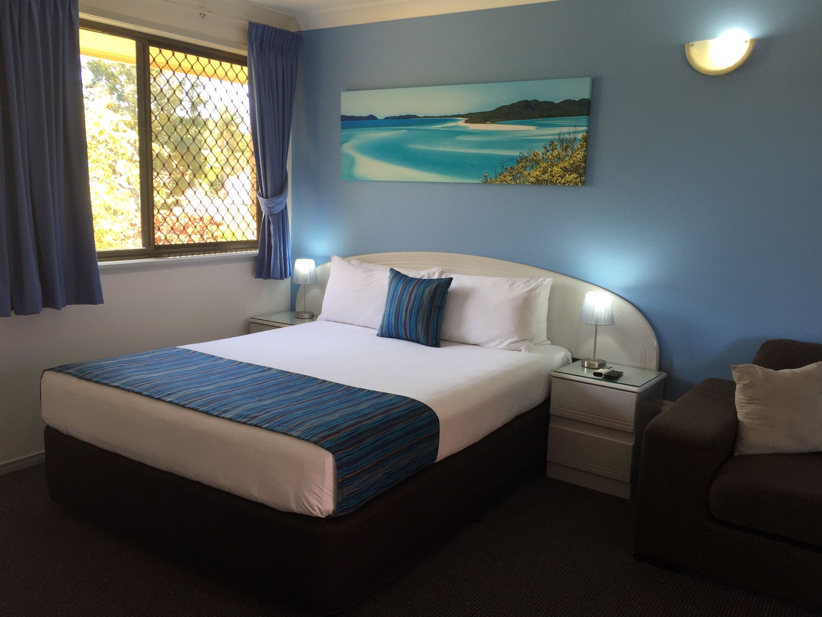 Una habitación de hotel con una cama grande, dos mesitas de noche, una silla, una ventana con cortinas y un cuadro en la pared.