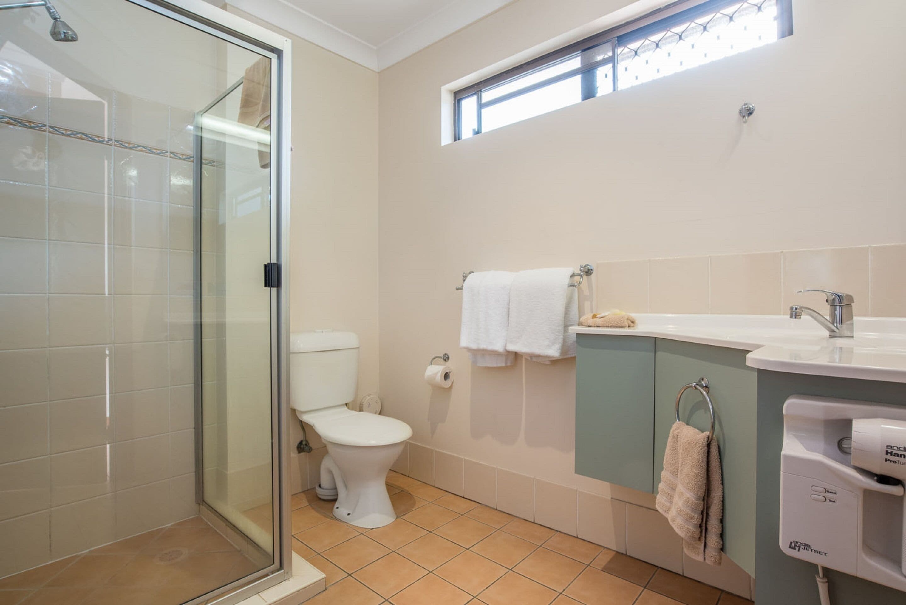 Un baño con cabina de ducha de vidrio, inodoro, lavamanos y ventana sobre el lavamanos.
