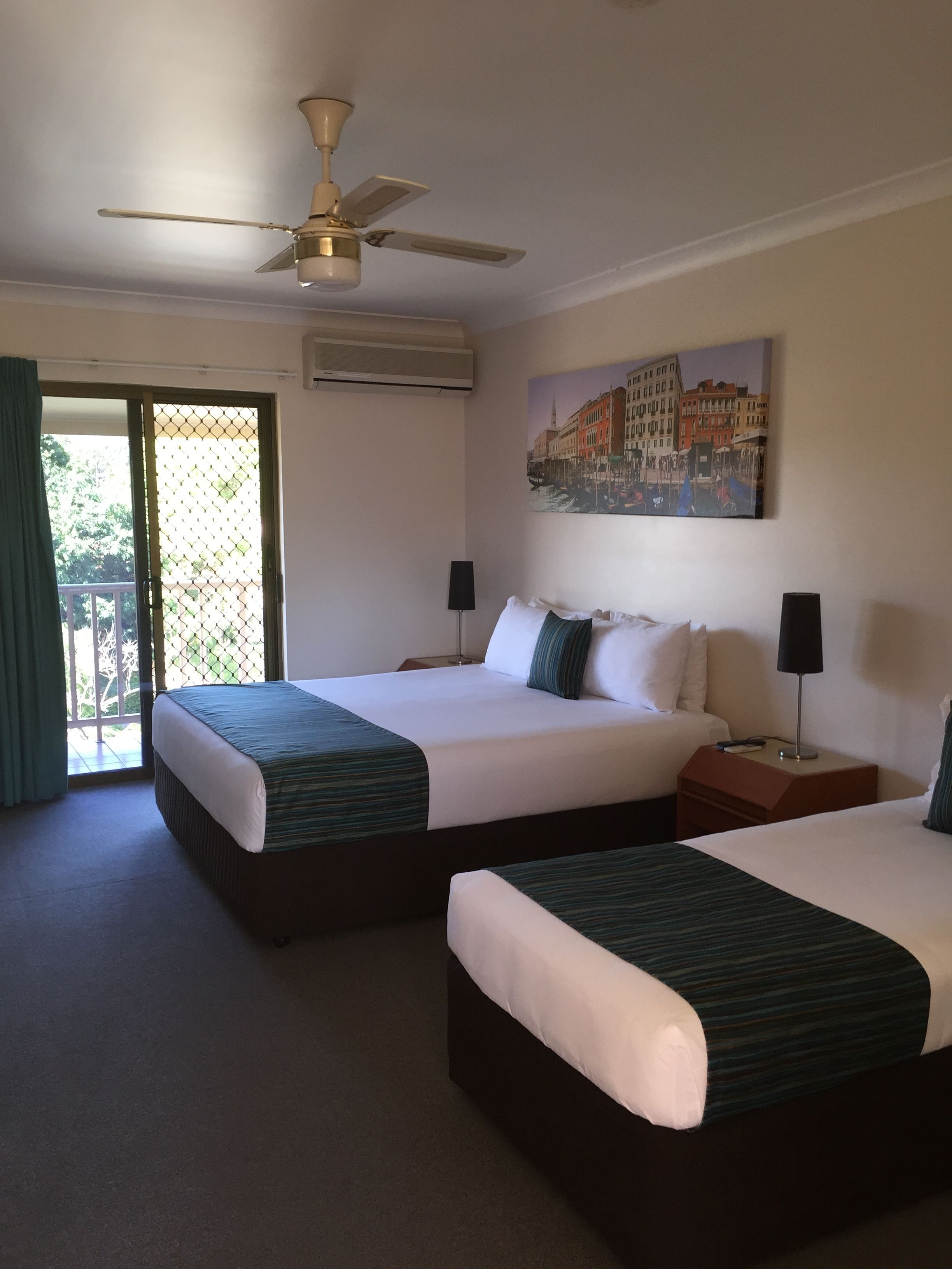 A hotel room with two beds, a ceiling fan, an air conditioning unit, a painting of a cityscape, and a sliding glass door leading to an outdoor area.
