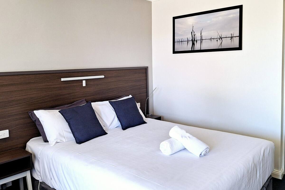 Una cama bien hecha con almohadas blancas y azul oscuro, una colcha blanca y una toalla enrollada sobre la cama.