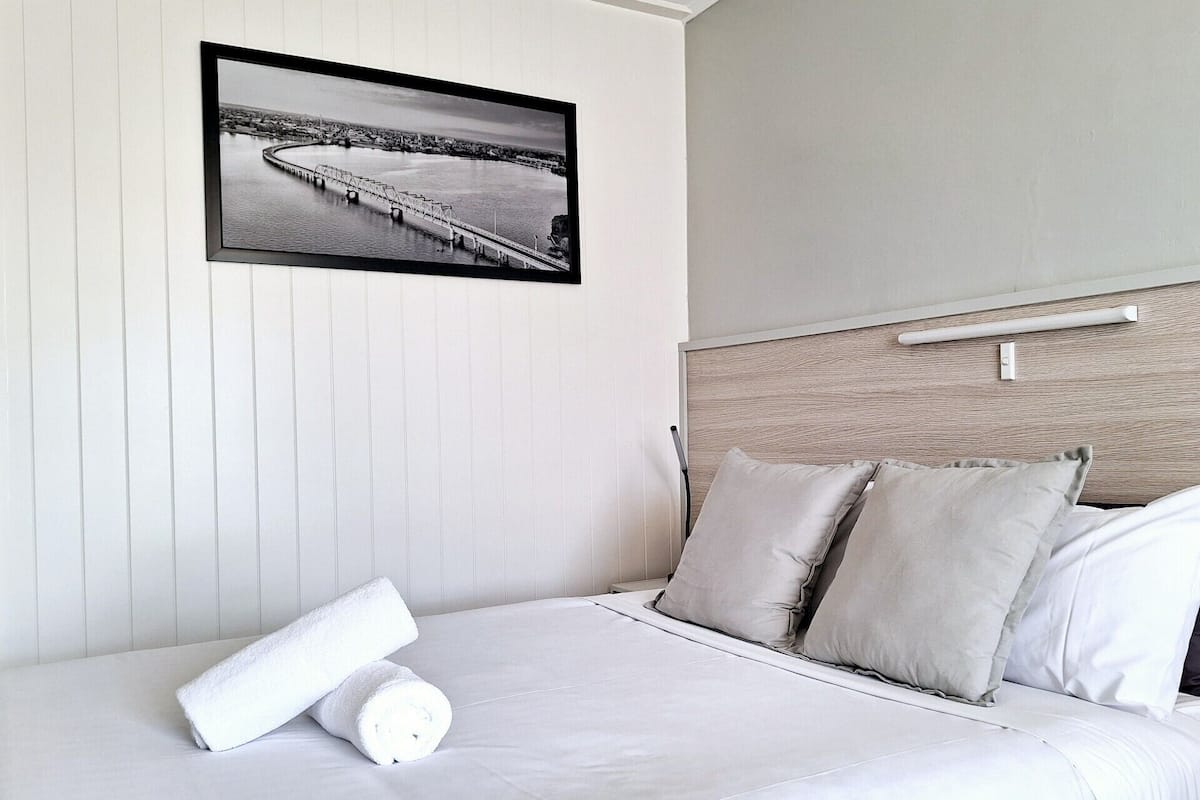 Una cama impecablemente hecha con sábanas y almohadas blancas, un cuadro de un puente en la pared y un cabecero de madera.