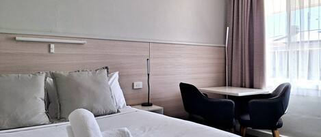 A hotel room with a neatly made bed, a desk with a chair, and a large window with curtains.
