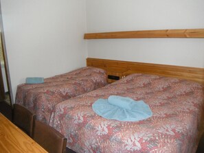 Una habitación pequeña y sencilla con dos camas individuales, cabecero de madera y pared blanca lisa.