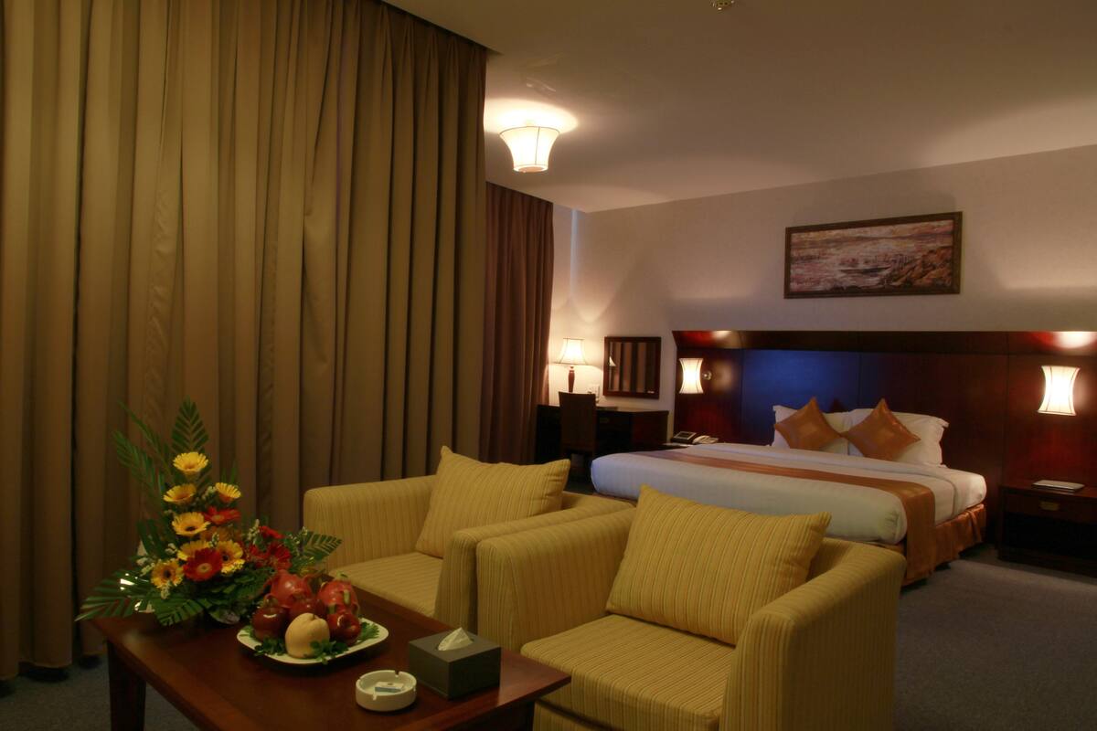 A hotel room with a bed, two armchairs, a table with a vase of flowers, and a painting on the wall.