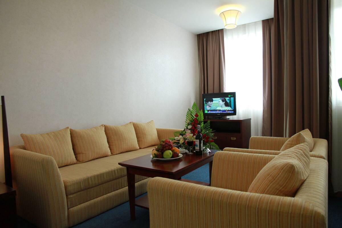 A hotel room with a sofa, armchair, and a small table with a fruit bowl and flowers. A television is on a stand against the wall.