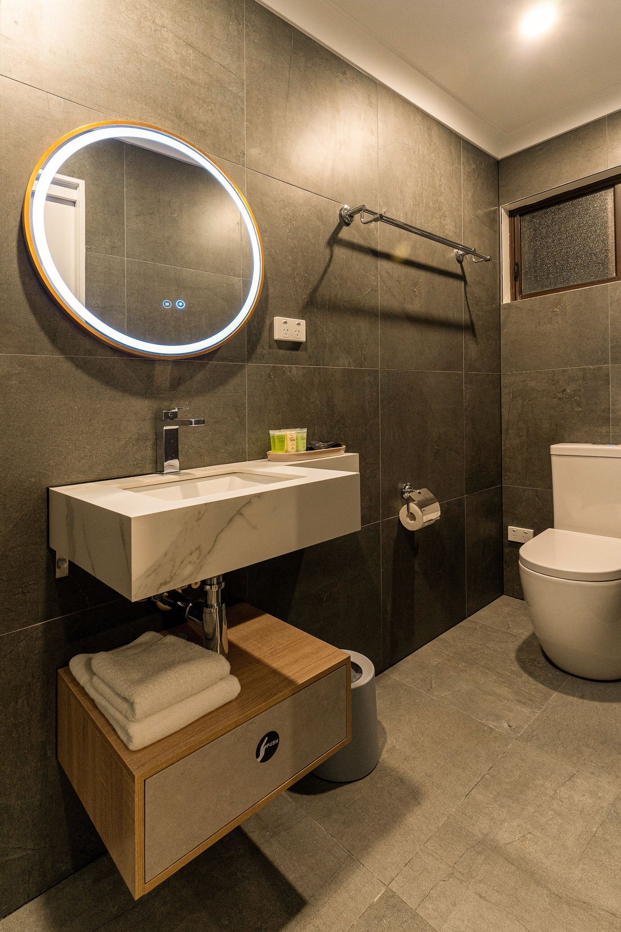 A modern bathroom with a marble sink, a round mirror with LED lighting, a towel rack, and a toilet.