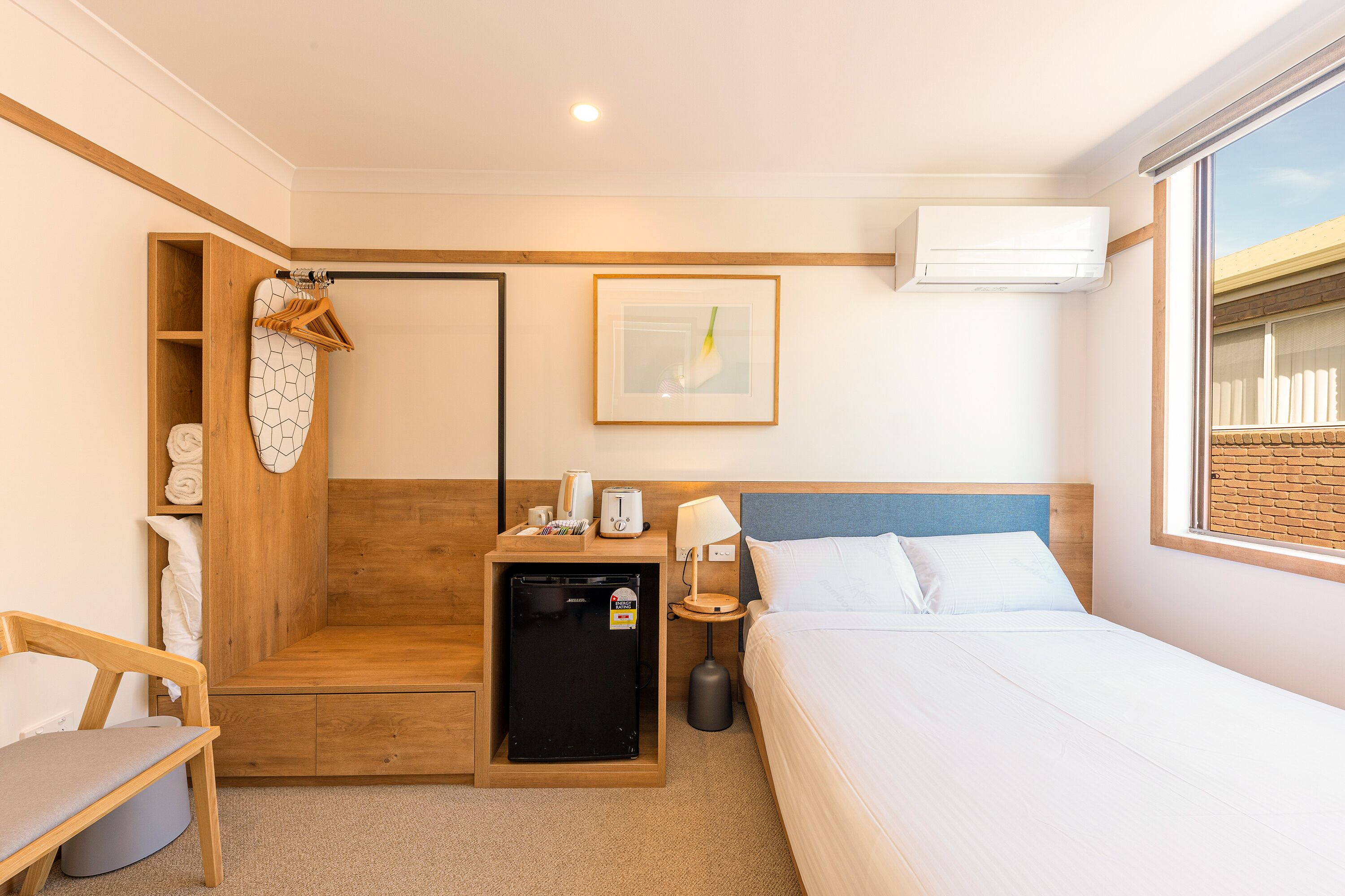 Habitación de hotel con cama, un pequeño escritorio de madera con refrigerador, zona de estar con una silla, perchero y un cuadro en la pared.