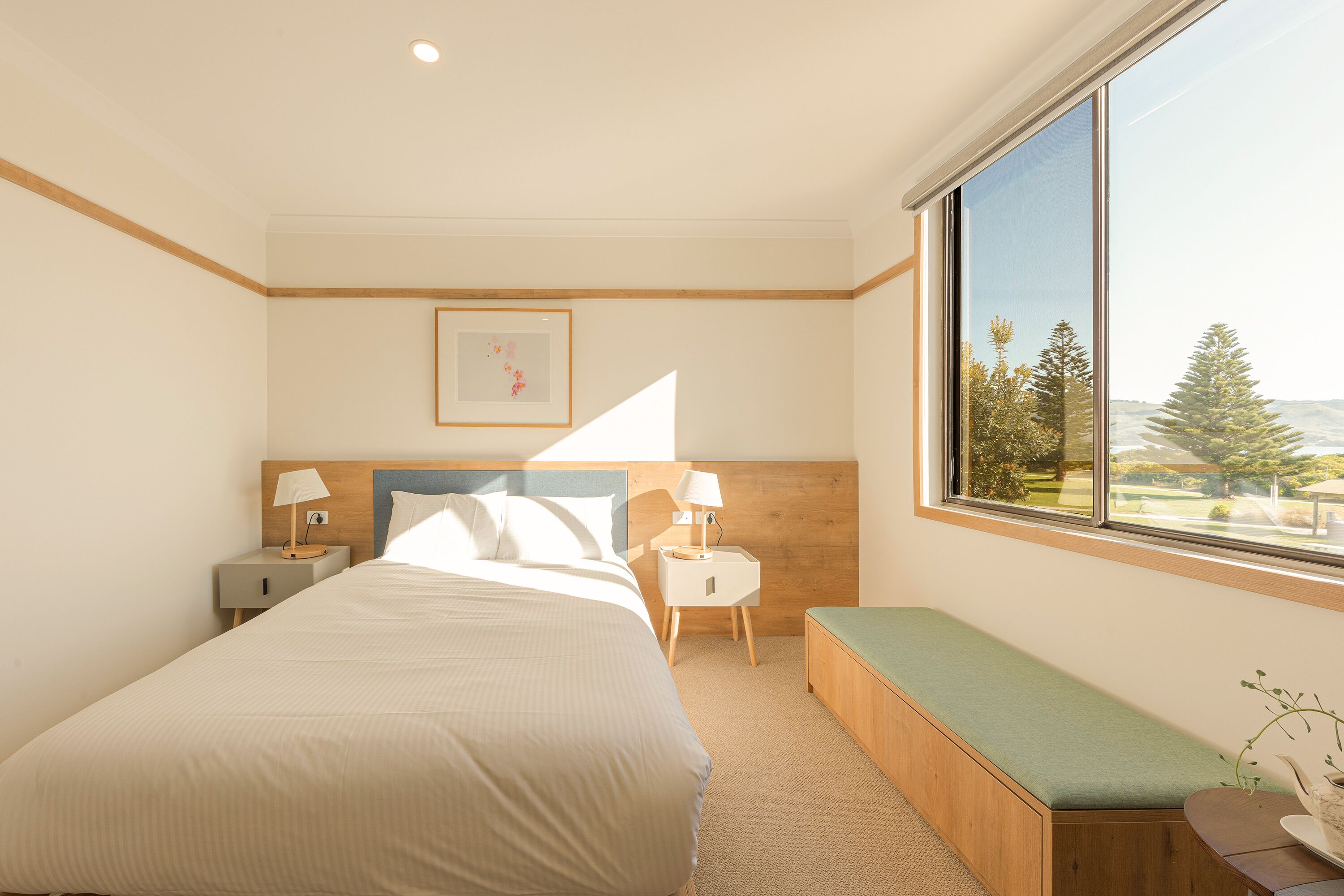 A hotel room with a large bed, a bench, a nightstand with a lamp, a framed picture on the wall, and a window with a view of trees.