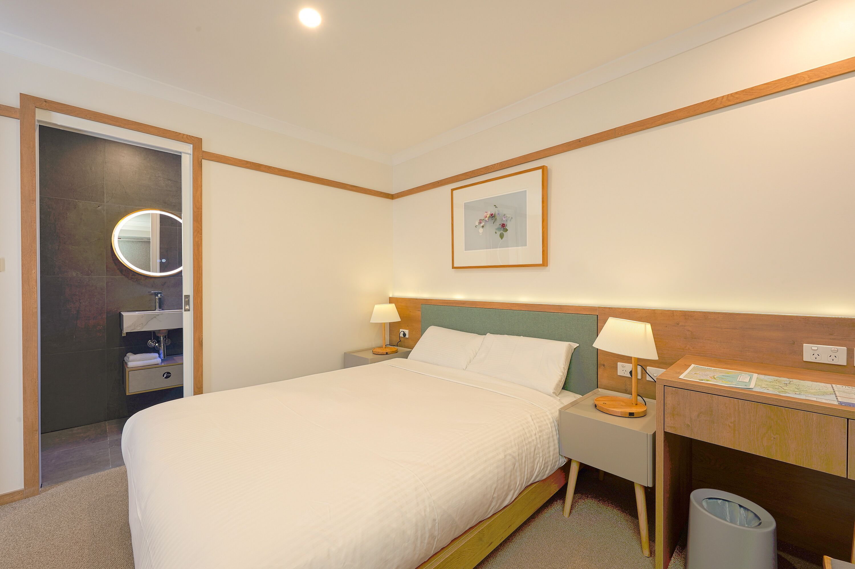 Habitación de hotel con una cama grande, mesitas de noche con lámparas, cabecera de madera, un escritorio pequeño con un mapa, un cesto de basura y un baño con espejo y toallero.
