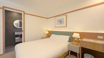 Habitación de hotel con una cama grande, mesitas de noche con lámparas, cabecera de madera, un escritorio pequeño con un mapa, un cesto de basura y un baño con espejo y toallero.