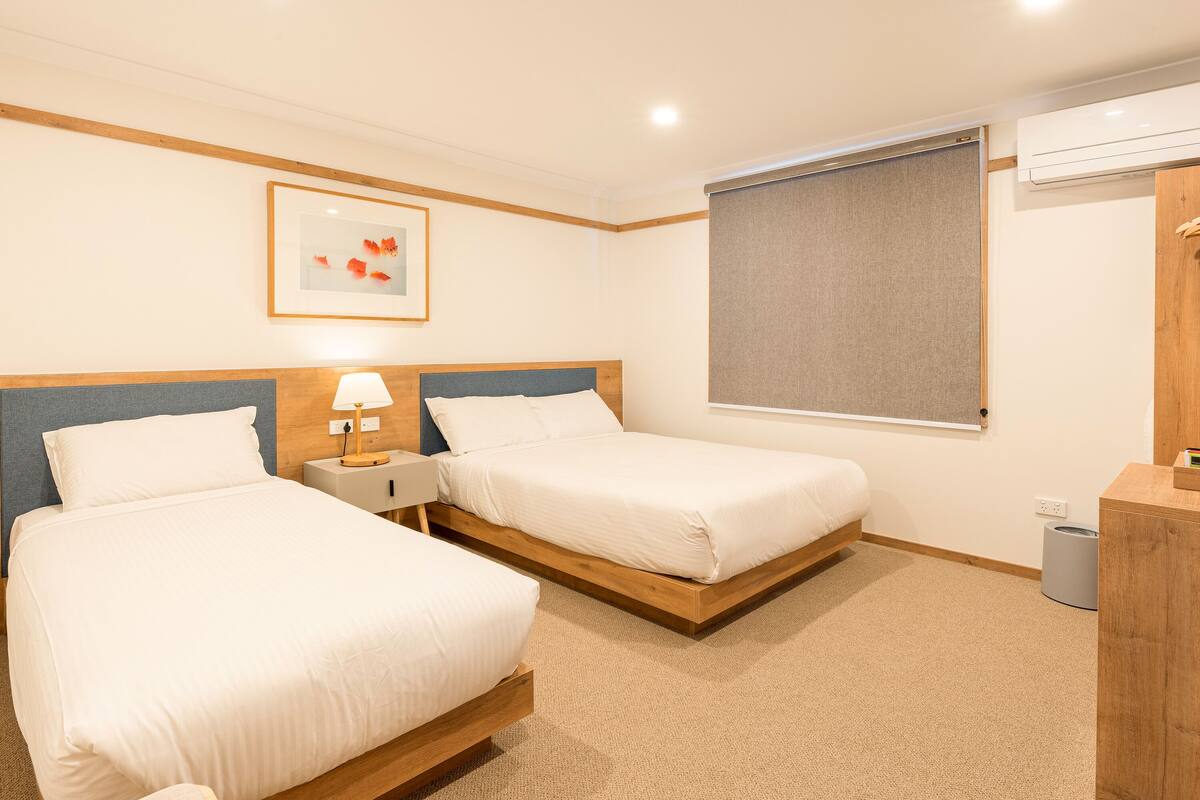 Habitación de hotel con dos camas individuales, una mesita de noche con lámpara, un cuadro enmarcado en la pared, una ventana con persiana y un aparador de madera.