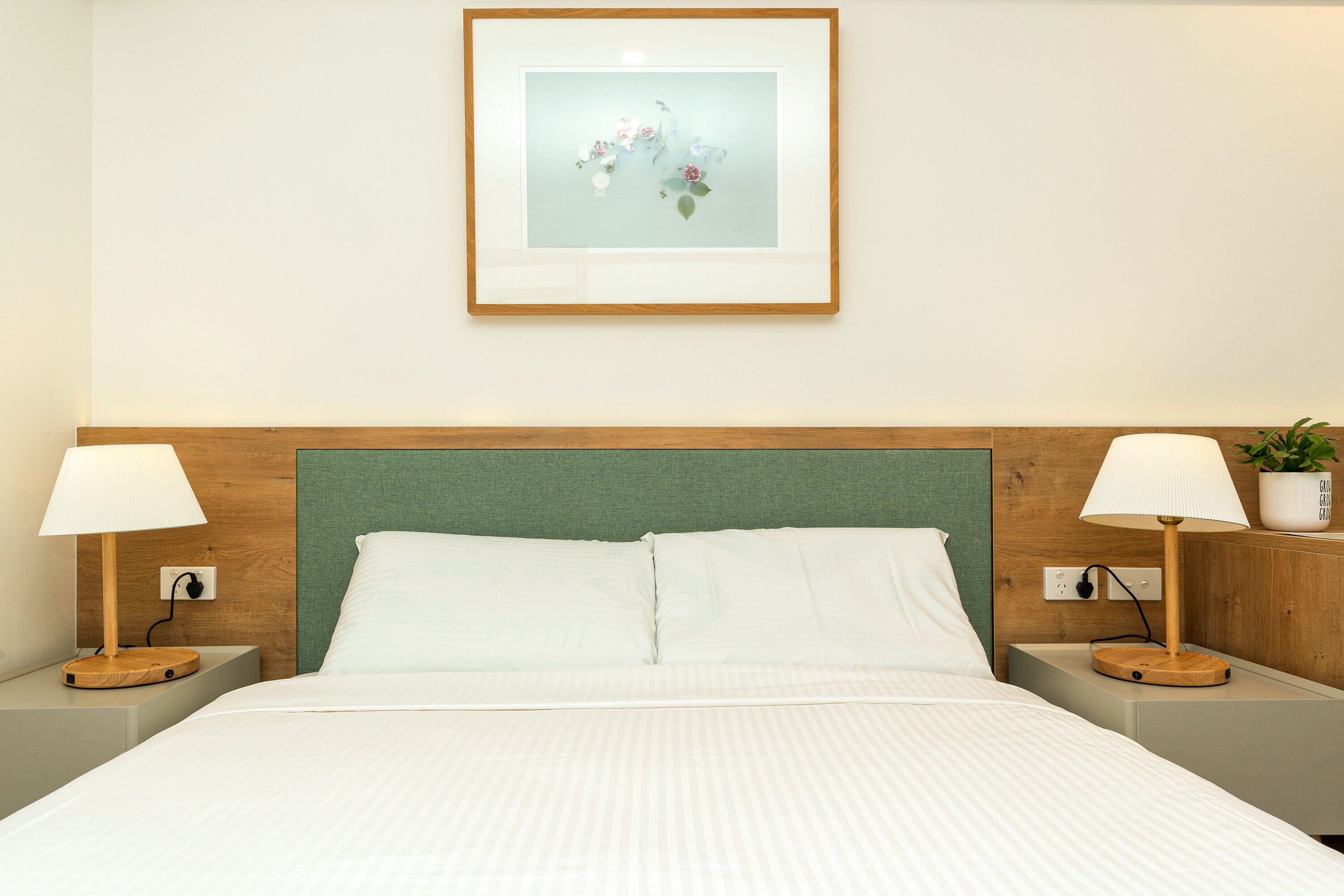 A neatly made bed with white linens, a green headboard, and two bedside tables with lamps and a potted plant.