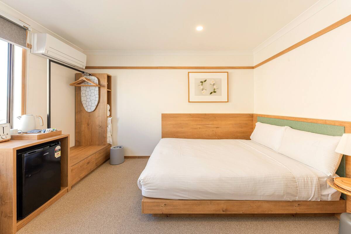 Habitación de hotel con una cama de madera, un escritorio pequeño con refrigerador, zona de ducha y un cuadro enmarcado en la pared.