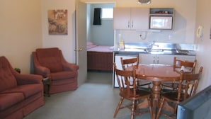 A compact living space with a red sofa, a wooden dining table with chairs, a kitchenette, and a small bed. - Blue Dolphin Motel (Timaru)