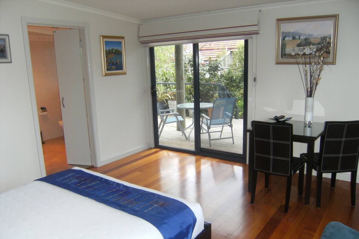Un dormitorio con cama, escritorio y silla, un balcón con mesa y sillas, y un cuadro en la pared.