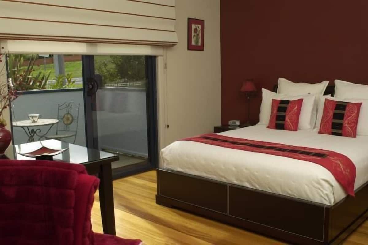 Un dormitorio con una cama, una silla roja, una mesita y vistas a través de un ventanal.