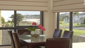 Un comedor con una mesa y sillas, un florero con flores, y vistas a una playa y casas a través de amplios ventanales.