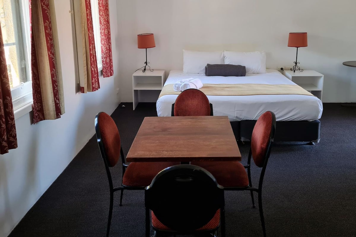 A hotel room with a bed, two bedside tables with lamps, a wooden table with four chairs, and red curtains.