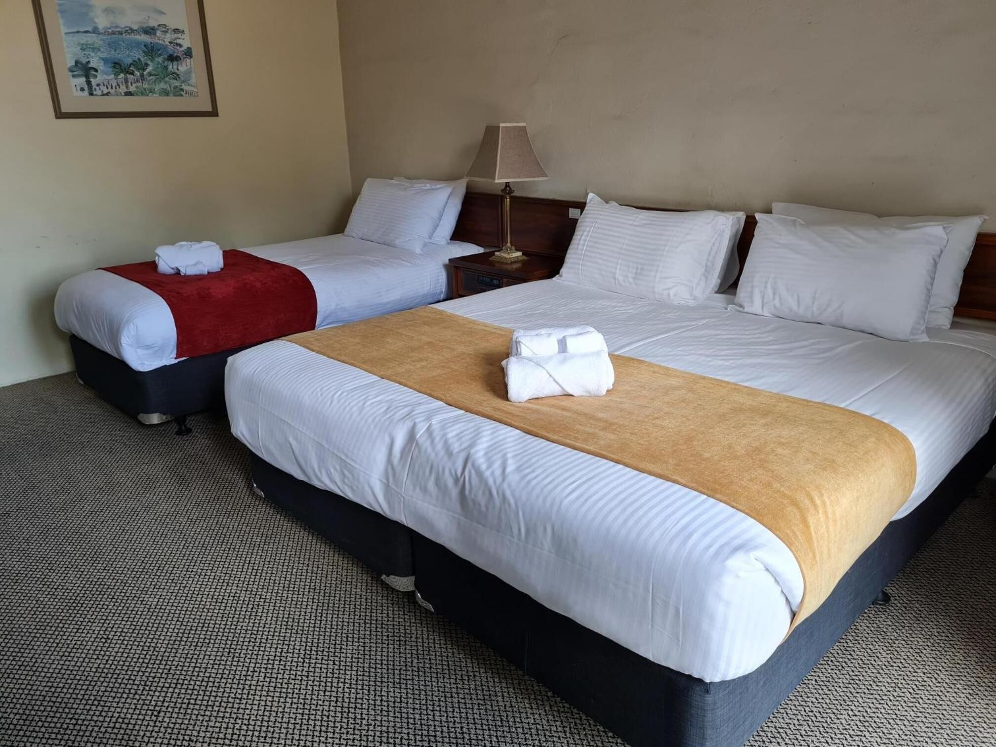 A hotel room with two beds, a red throw blanket on the left bed, a brown throw blanket on the right bed, and a lamp on the nightstand.