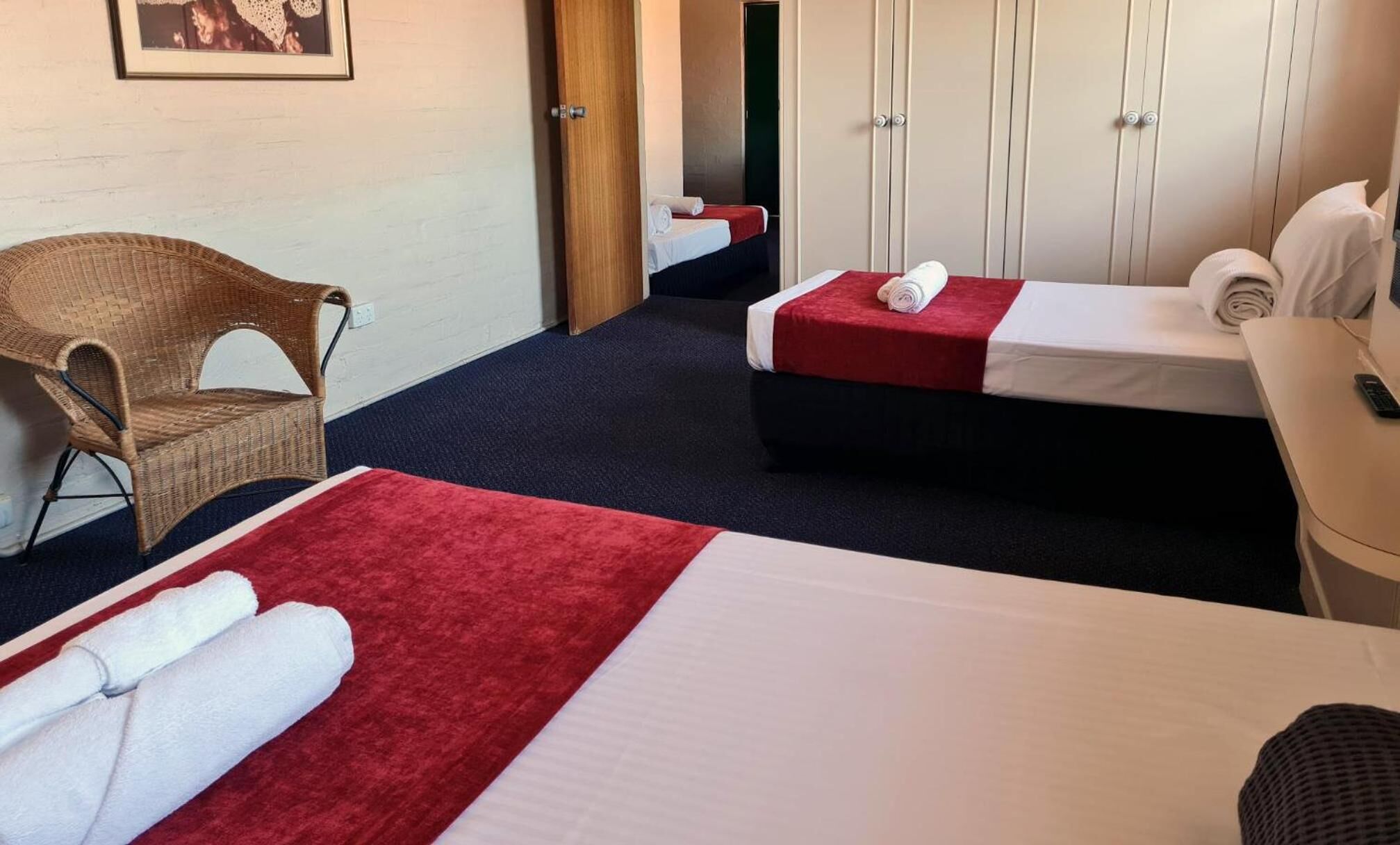 Une chambre d’hôtel avec deux lits, une chaise en osier, une armoire et un tableau encadré au mur.