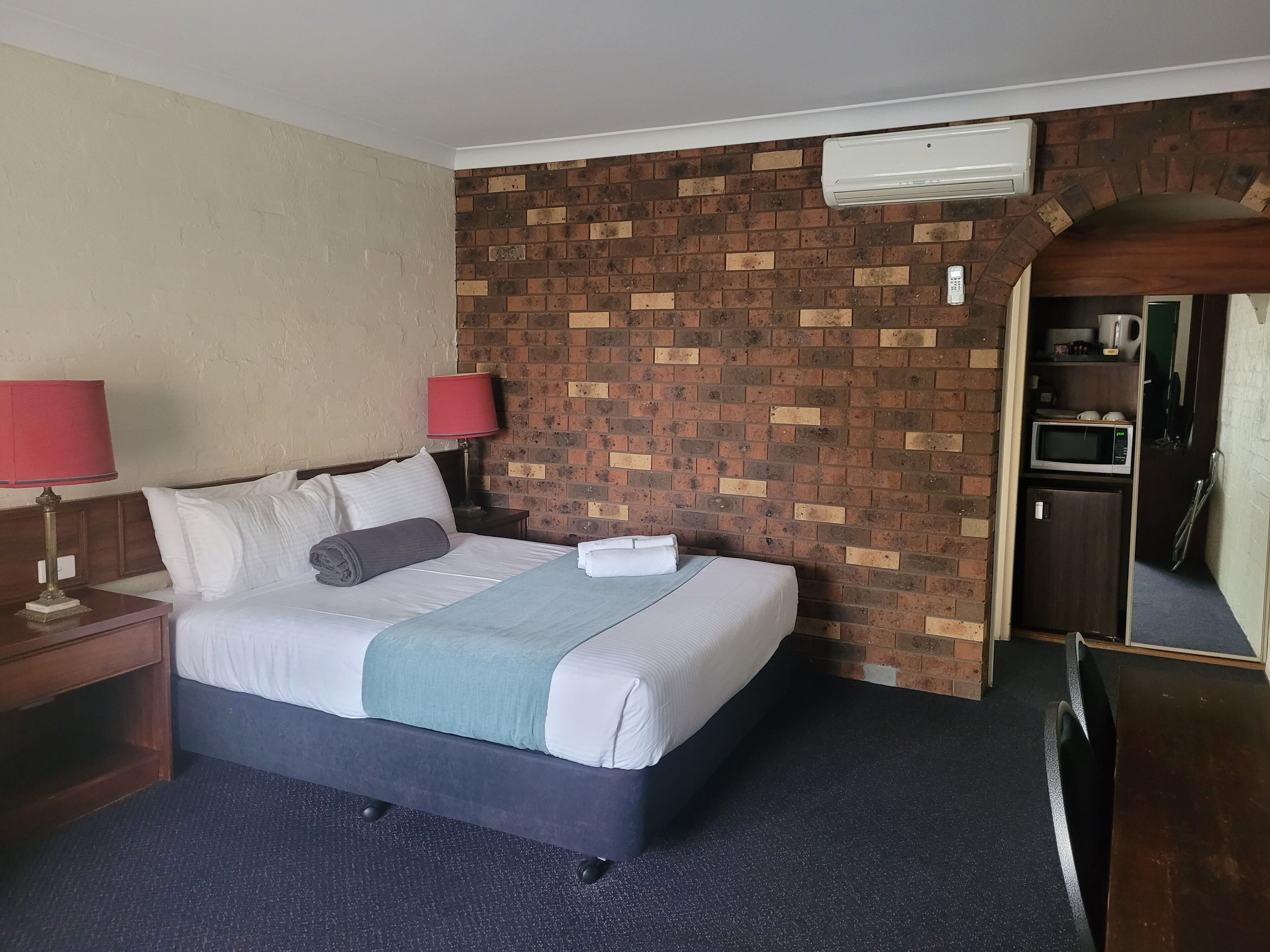 Habitación de hotel con cama, dos lámparas de noche, pared con diseño de ladrillo, unidad de aire acondicionado y microondas.