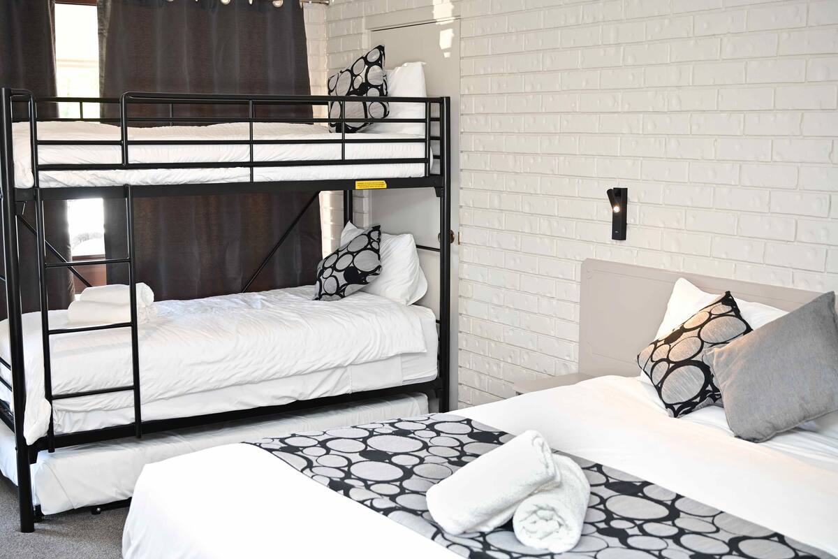 Een slaapkamer met stapelbedden, witte beddengoed en gestreepte kussens, een witte baksteenmuur en een raam met gordijnen.