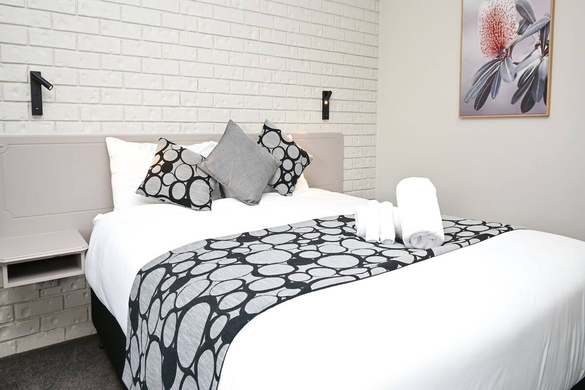 Een net opgemaakt bed met een geborreld sprei, kussens en een opgerolde handdoek tegen een witte bakstenen muur.