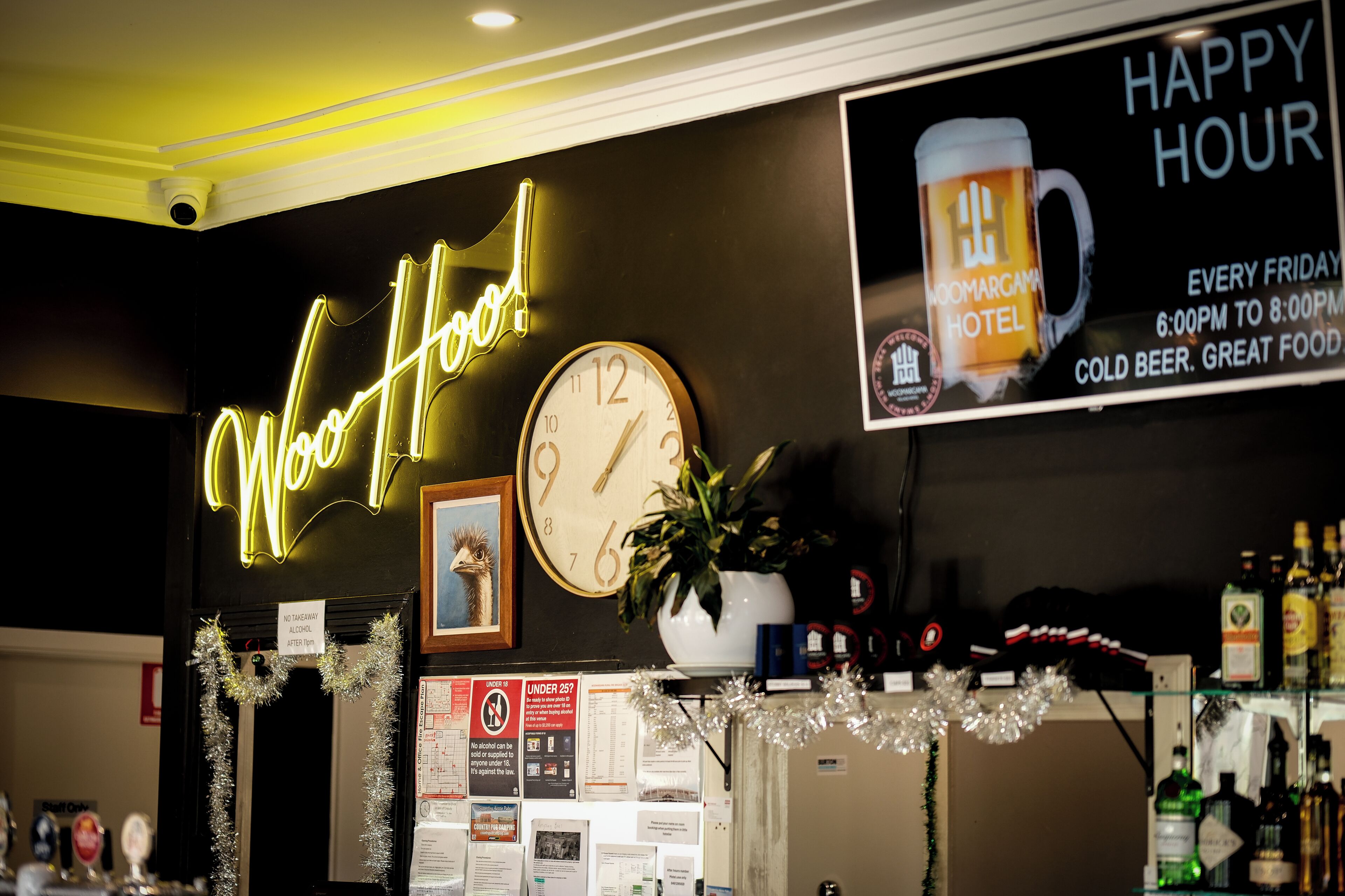 A hotel reception area with a neon 'Wow! Wow!' sign, a clock, a 'Happy Hour' poster, and a plant on the counter.