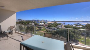 Un balcón con mesa y sillas, con vista a una zona costera con edificios, una playa y una extensión de agua.