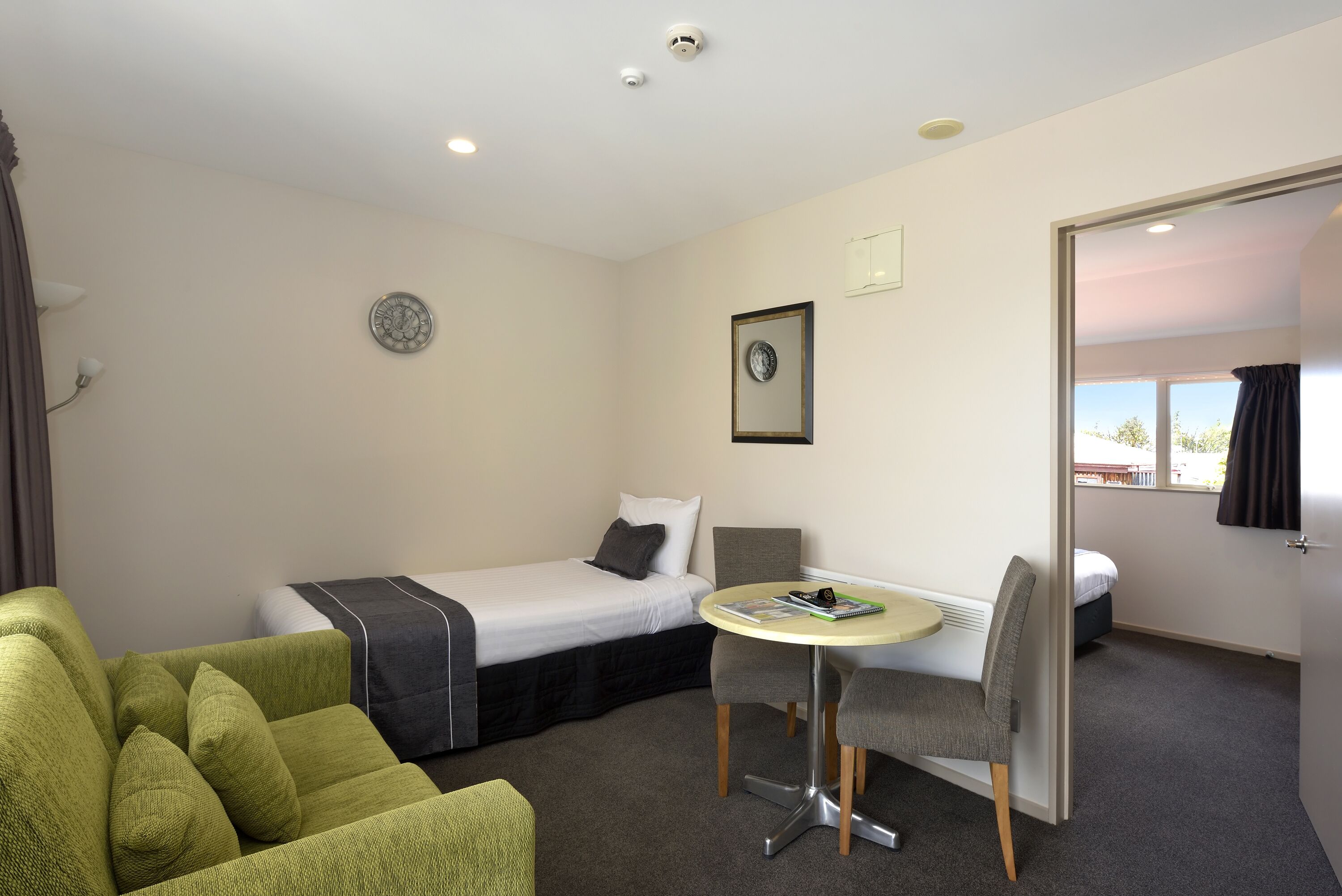 Una habitación de hotel con una cama, un sofá verde, una mesita con una silla y una puerta que da a otra habitación.