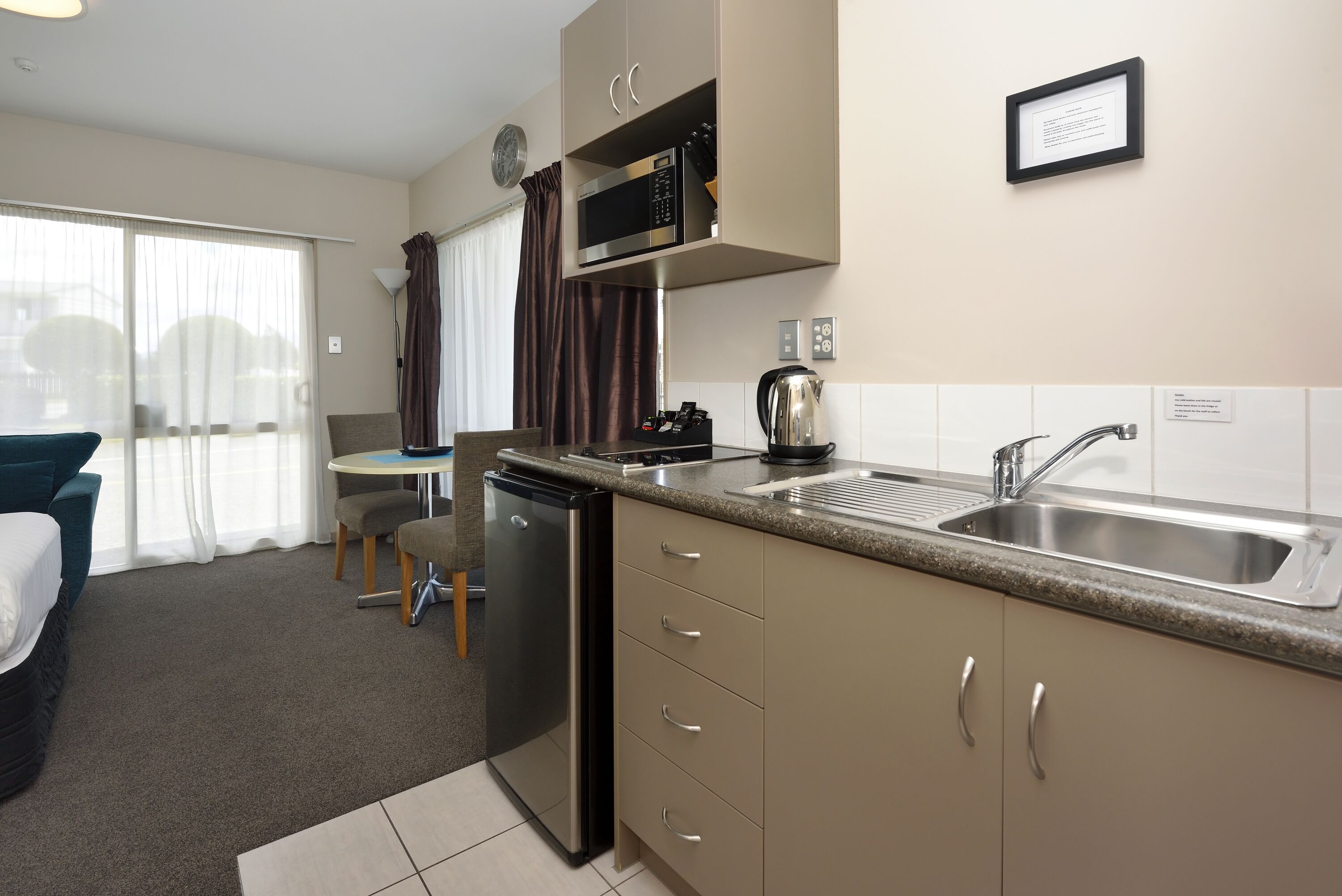 A compact hotel kitchen with a dishwasher, microwave, and sink. There is a small dining area with a table and chairs, and a bed in the adjacent room.