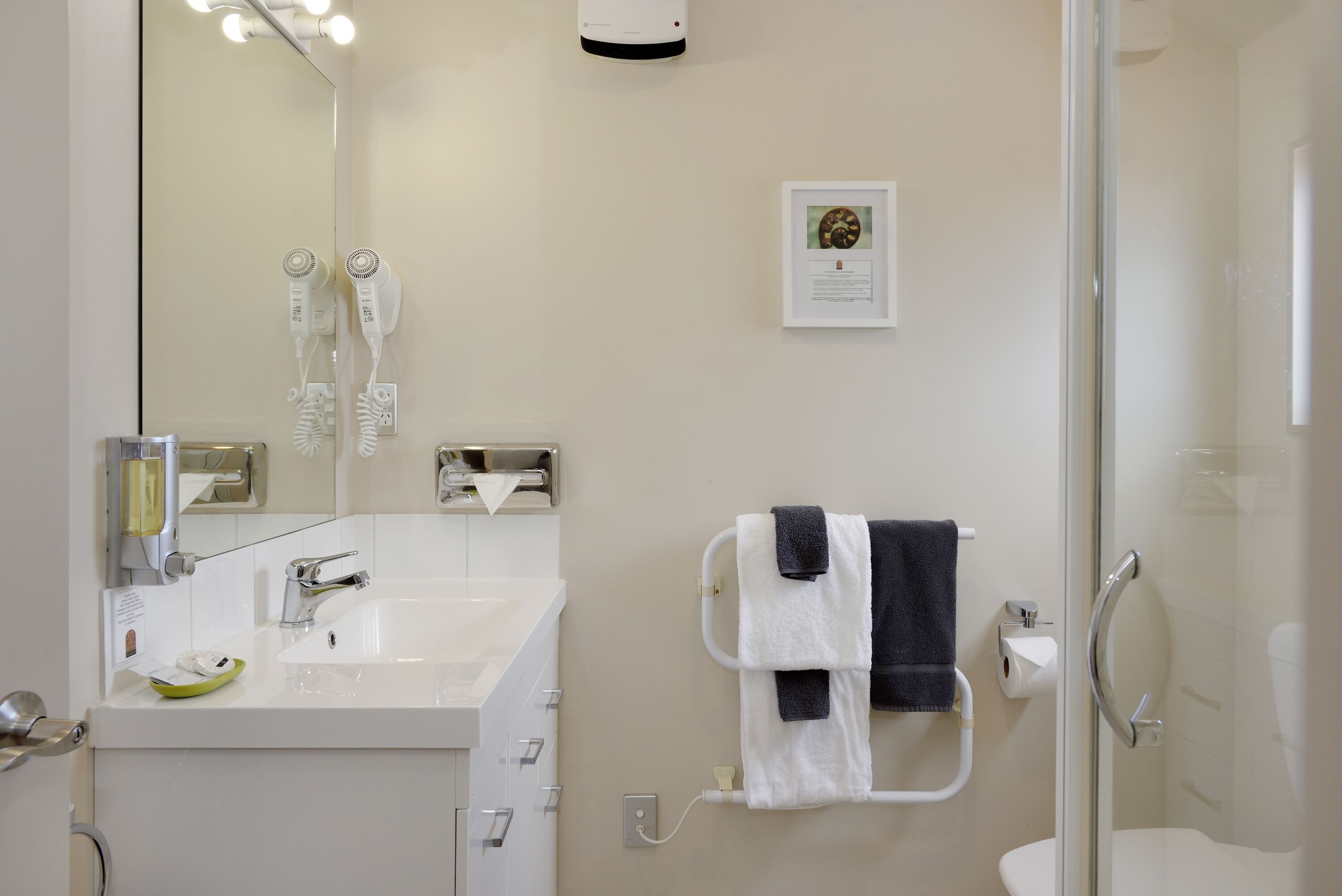 Un baño moderno con un lavamanos blanco, un espejo, dos secadores de pelo, un perchero con toallas e inmenso box de ducha con mampara de cristal.