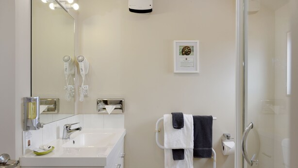 A modern bathroom with a white sink, a mirror, two hair dryers, a towel rack with towels, and a glass shower enclosure. - 171 On High Motel (Blenheim)