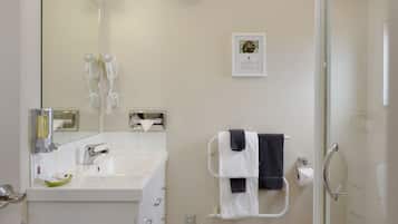 Un baño moderno con un lavamanos blanco, un espejo, dos secadores de pelo, un perchero con toallas e inmenso box de ducha con mampara de cristal.