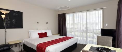 Une chambre d’hôtel avec un lit, un bureau, une chaise, une lampe et une grande fenêtre avec des rideaux.