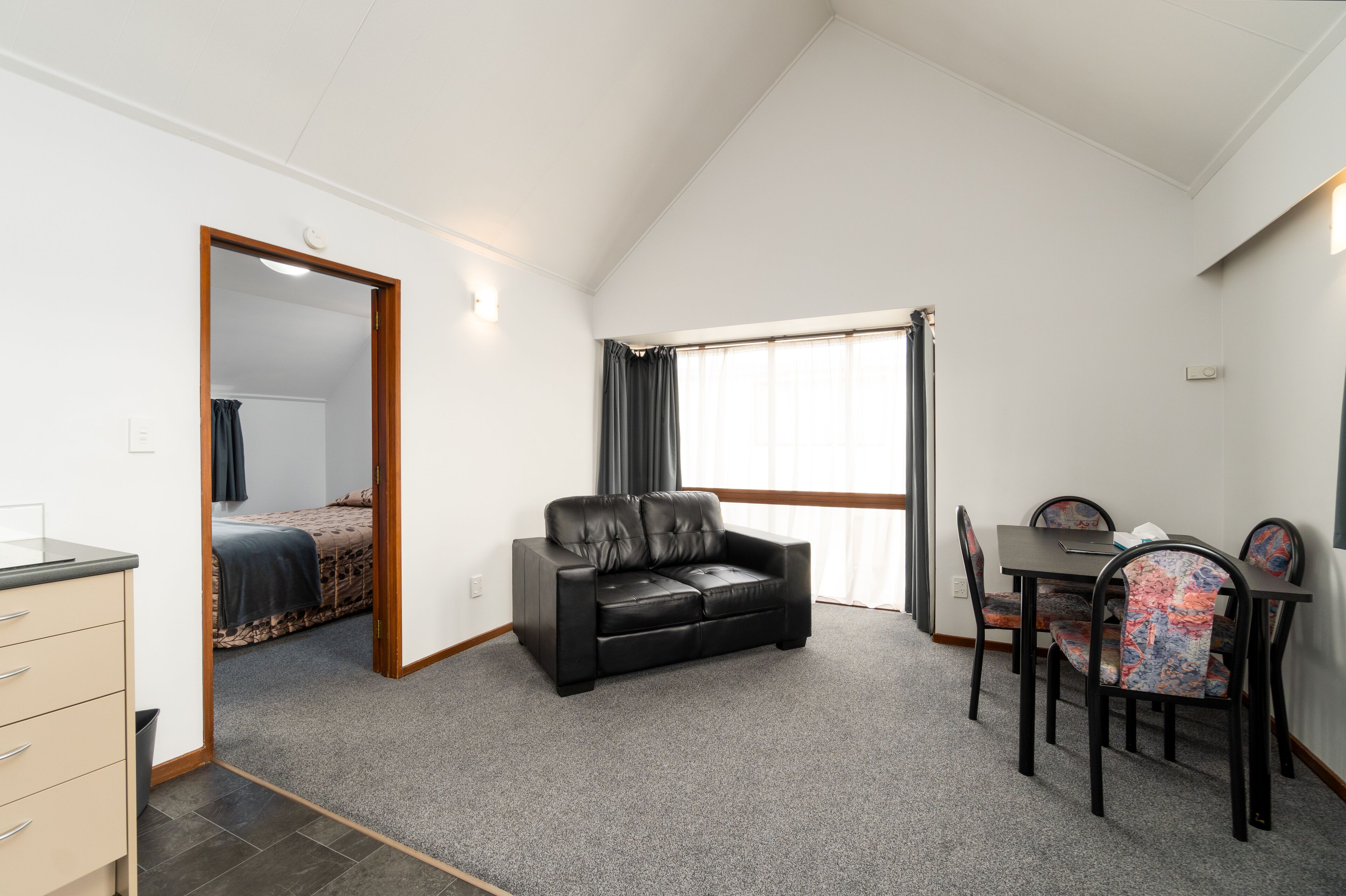 A room with a black leather sofa, a dining table with four chairs, a small kitchenette, and a large window with curtains.
