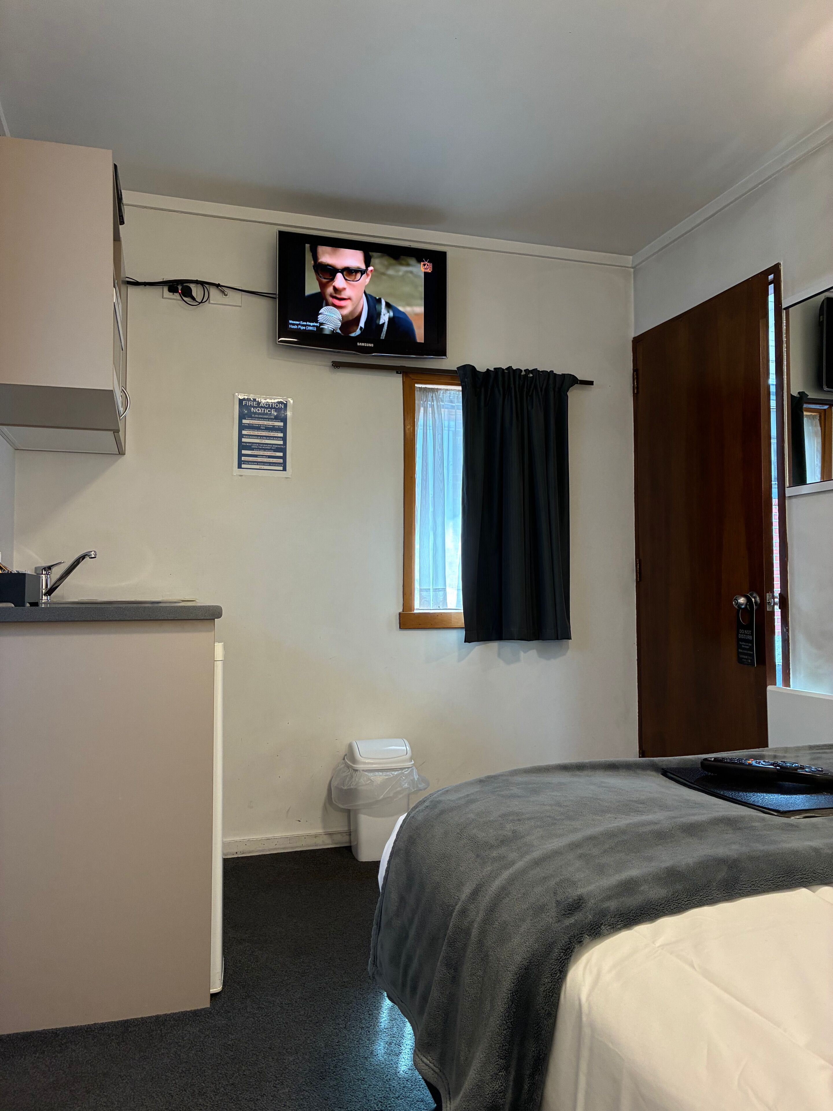 A hotel room with a bed, a desk, a television, a window with curtains, a door, and a notice on the wall.