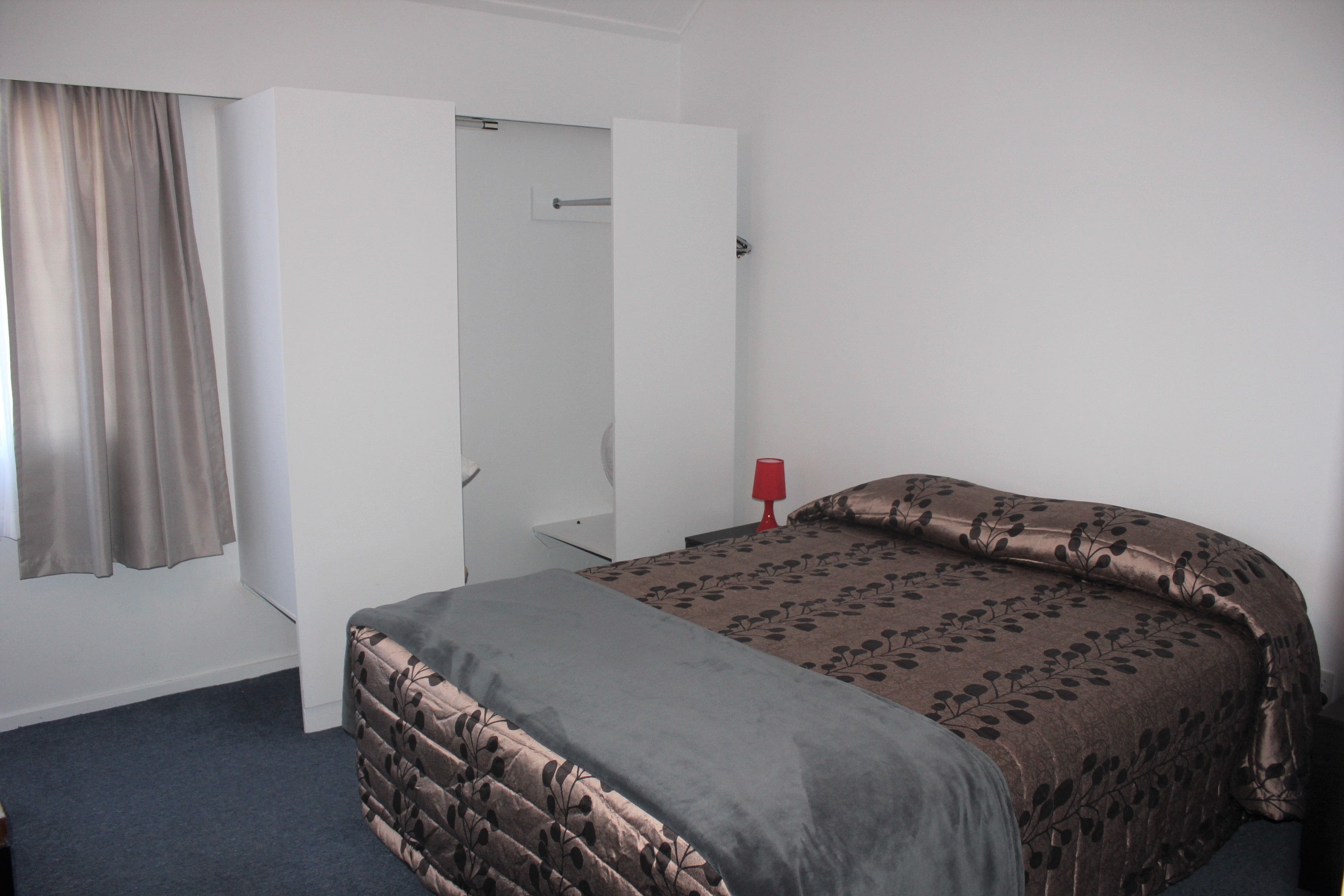 Um quarto com cama, guarda-roupa, luminária vermelha e janela com cortinas.