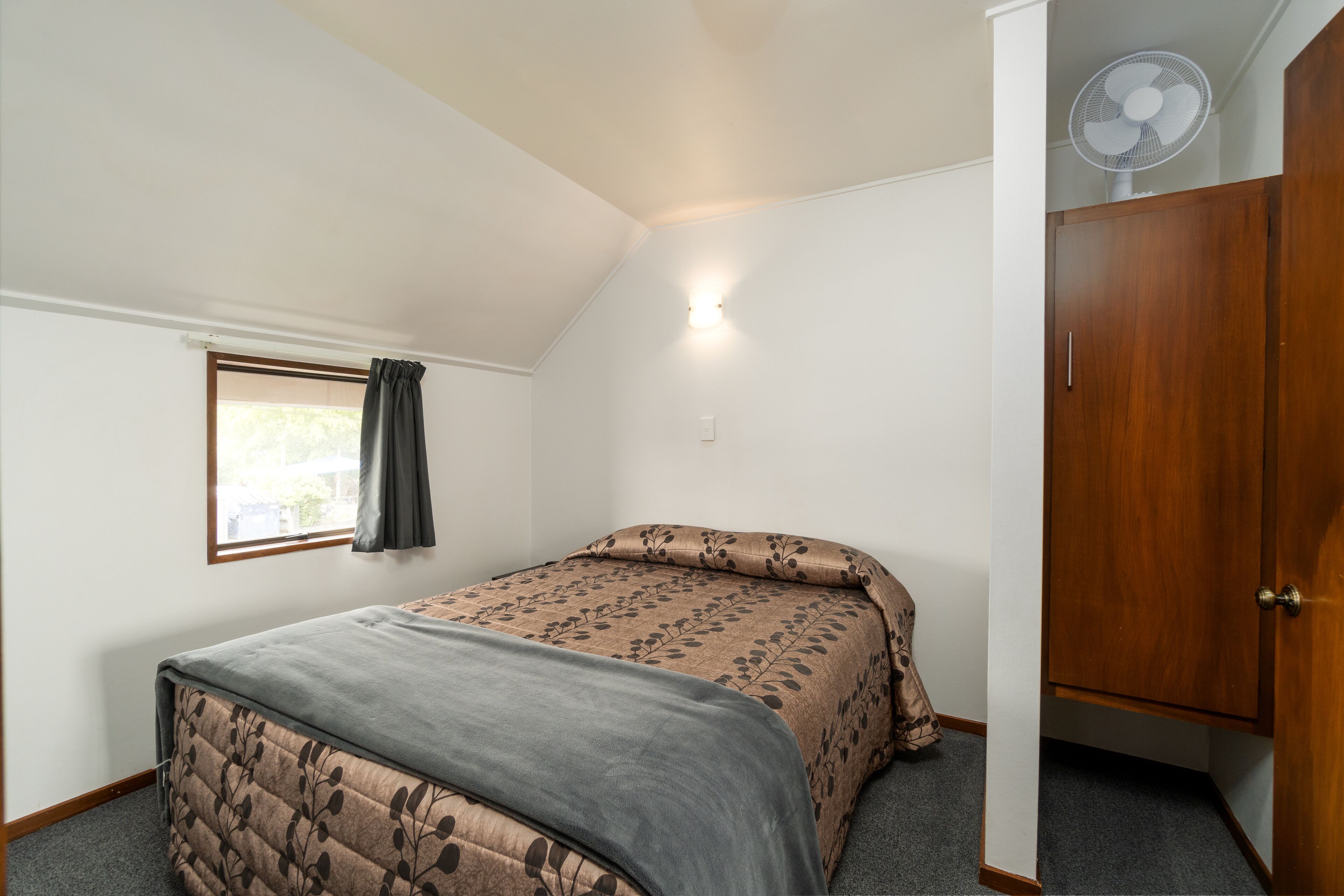 A bedroom with a bed, a window with a curtain, a wooden wardrobe, and a ceiling fan.
