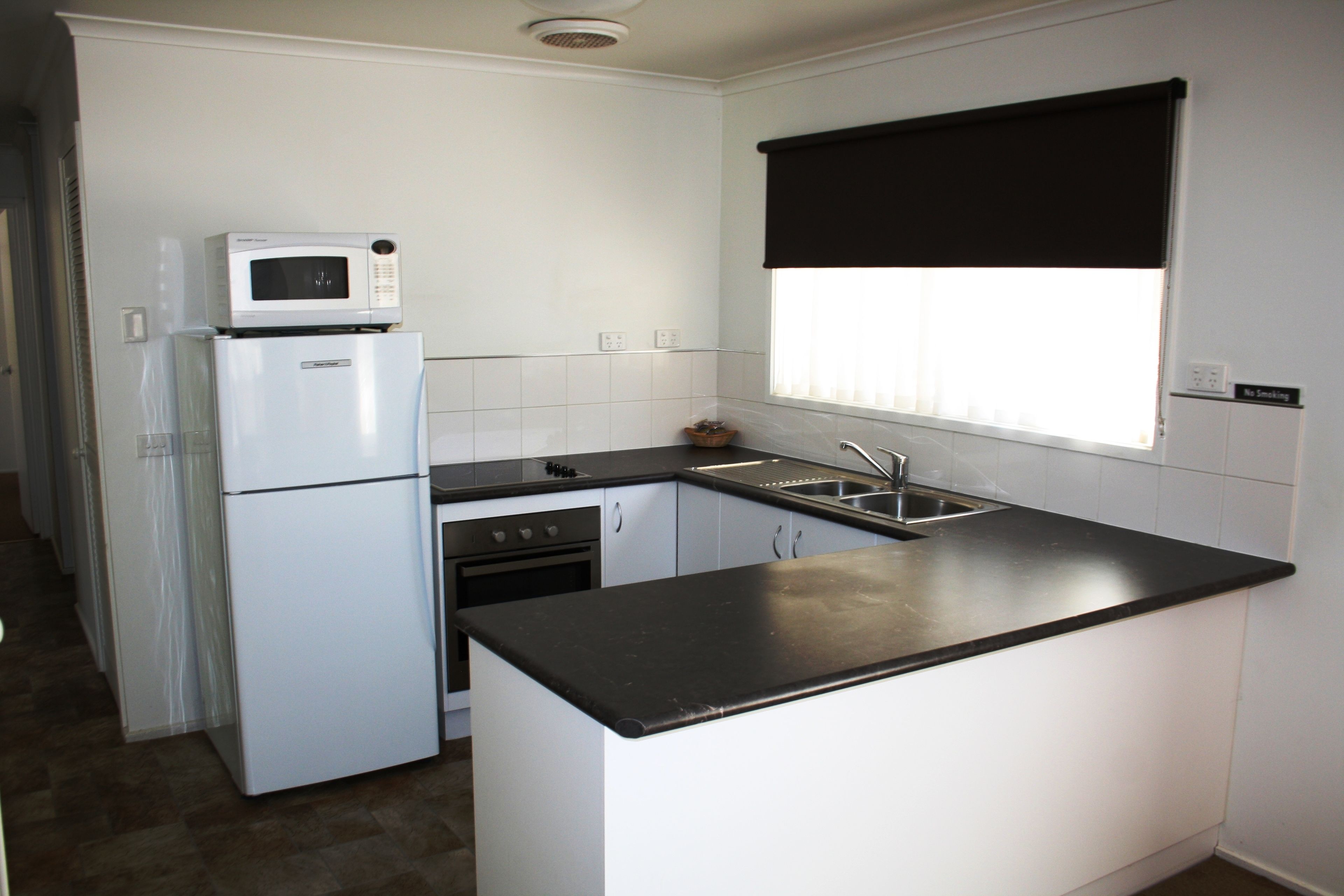 A compact kitchen with white cabinets, a black countertop, a microwave on the refrigerator, a sink, and a window with a black roller blind.