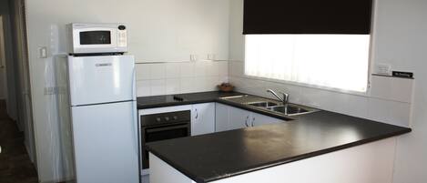 Cocina compacta con armarios blancos, encimera negra, microondas sobre el refrigerador, un lavabo y una ventana con persiana enrollable negra.