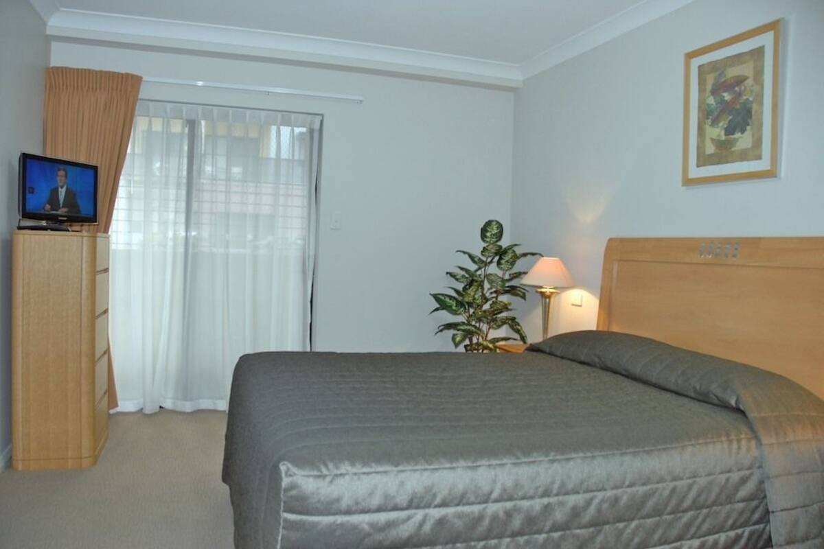 A hotel room with a bed, a television on a wooden cabinet, a plant, and a painting on the wall.
