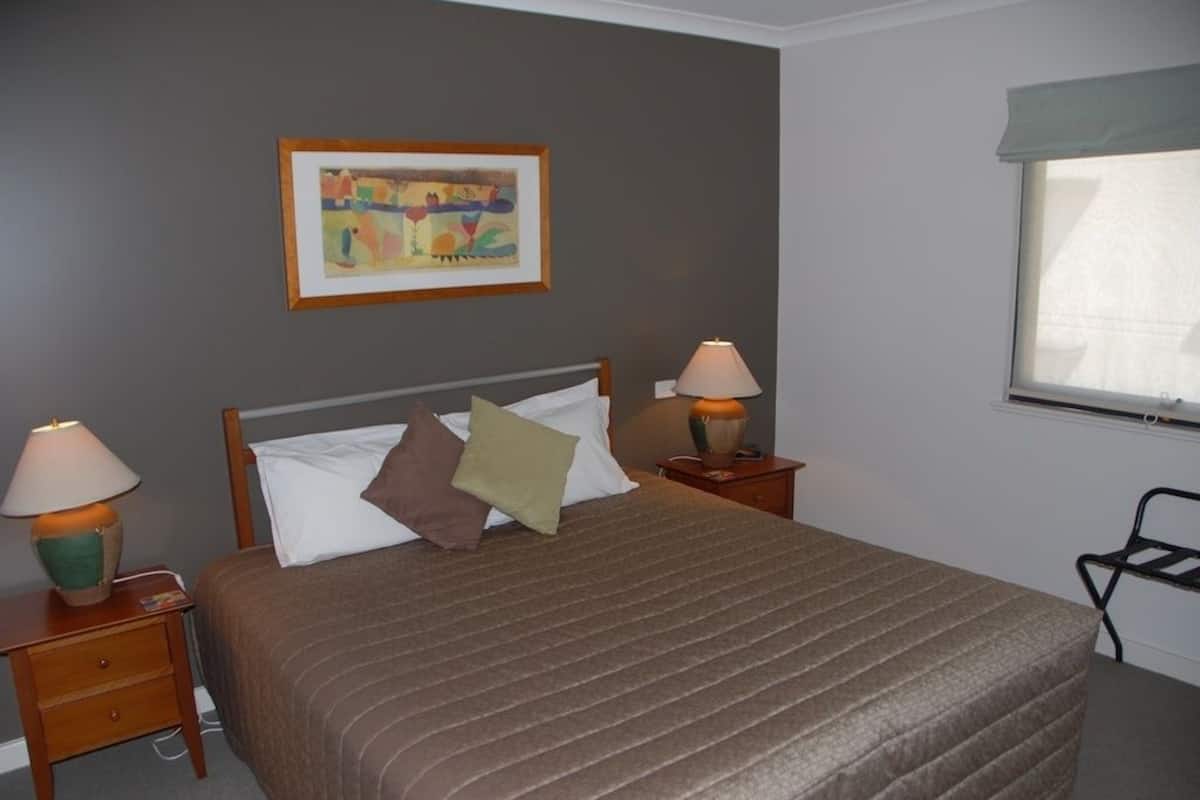 A neatly made bed with a brown bedspread, two bedside lamps, a framed abstract painting on the wall, and a window with a white blind.