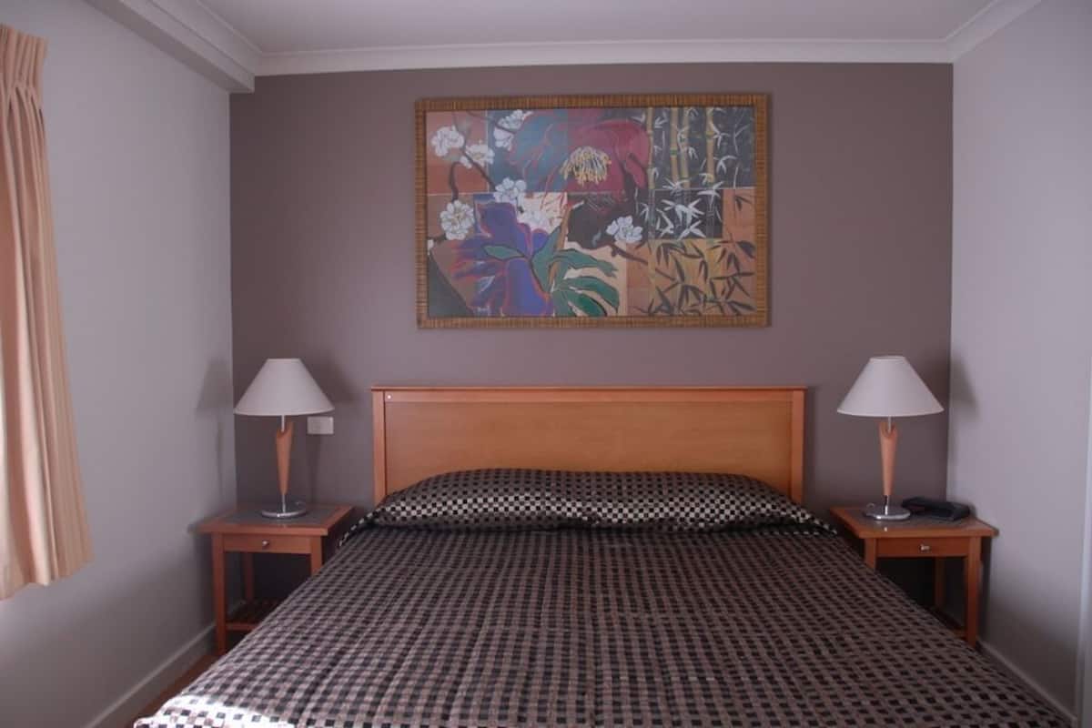 A hotel room with a checkered bedspread, two bedside lamps, a wooden headboard, and a floral artwork on the wall.