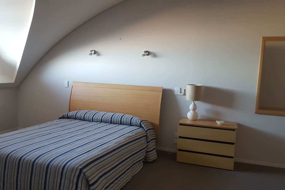 A bedroom with a sloped ceiling, a bed with striped bedding, a bedside table with a lamp, and a wall-mounted mirror.
