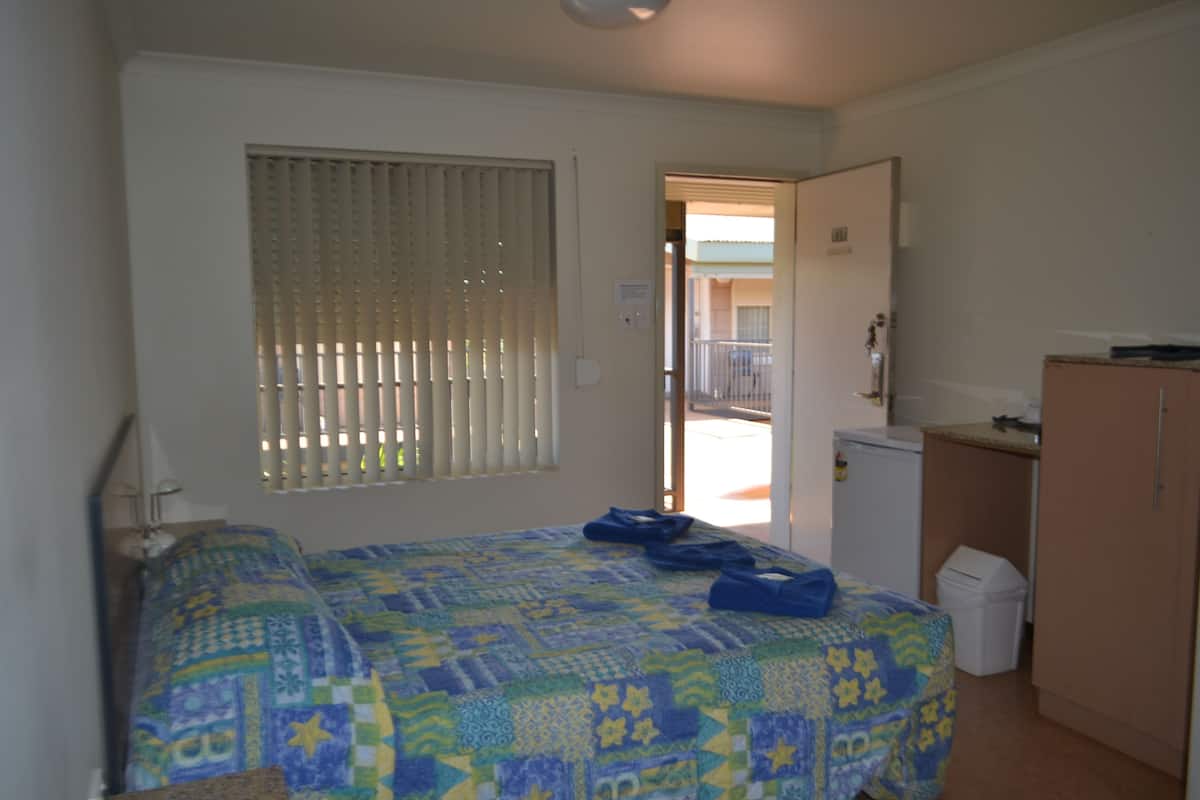 Ein kleiner Raum mit einem Bett, einem Fenster mit Jalousien, einer Tür zu einem Balkon und einer Küchenzeile.